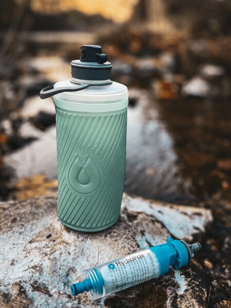 Сгъваема бутилка Flux™ HydraPak<sup>&reg;</sup> с поставен воден филтър HydraPak<sup>&reg;</sup> (28 мм) лежи на камък до поток. Снимка: Rigad