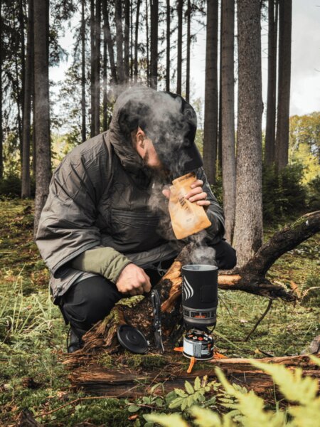 Мъж в изолирано яке коленичи в гората до котлон Jetboil, от който се издига пара. Приготвя топла храна или напитка като източник на енергия и топлина по време на есенното пътуване. Снимка: Rigad