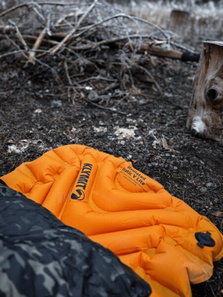Надуваема постелка Klymit Insulated Static V Lite в оранжев цвят на земята до огнището, частично покрита със спален чувал с камуфлажен десен. Изолация от студ и влажност при спане на открито. Снимка: Rigad