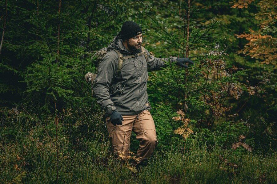 Човек в есенна гора в сиво зимно яке Yukon с изолация Climashield™ Apex® от Helikon-Tex®, шапка и ръкавици, с раница; демонстрация на правилно наслояване и защитен връхен слой за влага и вятър. Снимка: Rigad
