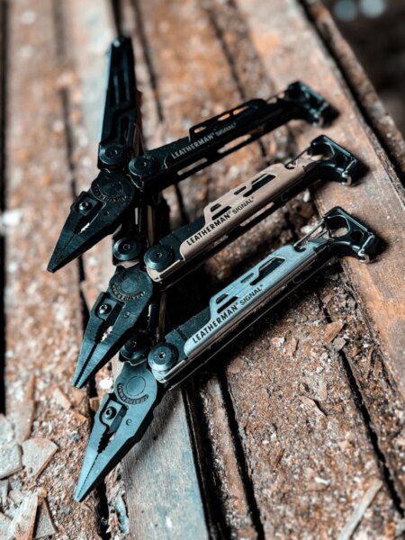 Три мултитула Leatherman Signal, които се различават само по цвят, поставени върху дървена повърхност.